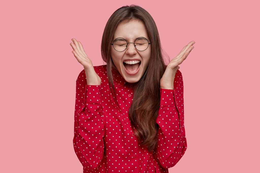 Positive Excited Woman Spreads Hands, Shouts With Happiness, Feels Excited And Overjoyed, Wears Fashionable Red Blouse, Models Against Pink Background. People, Human Emotions And Feelings Concept