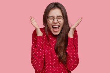 Positive Excited Woman Spreads Hands, Shouts With Happiness, Feels Excited And Overjoyed, Wears Fashionable Red Blouse, Models Against Pink Background. People, Human Emotions And Feelings Concept